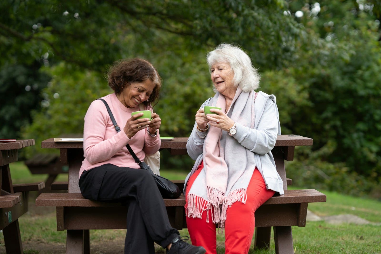 Two older women laughing over a cup of tea on a park bench